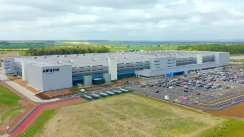 A sprawling grey box building, which is four storeys high, stands next to a car park. The building has an Amazon sign in the corner and is surrounded by fields.