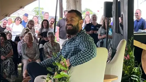 A man with short brown hair with shorter sides and a brown beard is smiling to the side. He is wearing a chequered blue shirt and sat on a beige seat on a stage. Behind him are several people sat in front of the stage on chairs and stood up at the back.