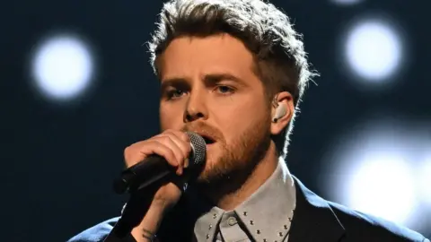 Singer-songwriter Alex Warren holds his microphone as he performs at the American Music Awards. Stage lights shine behind him.