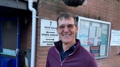 A photo of Jim Maltby. He is smiling to the camera outside the community centre. He is wearing a purple jumper with a navy top underneath. He has brown short hair and is wearing glasses.