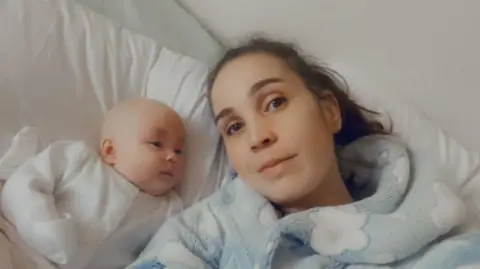 Natalie Natalie is lying on a bed beside a baby dressed in a white outfit. The person is wearing a light blue fleece robe with a cloud pattern, and both are positioned against white pillows and bedding.