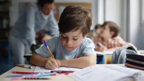 Getty Images A child with short brown hair writing in a book at a desk in school.