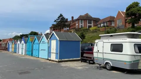 Alice Whaley/BBC A campervan parked next to beach huts in Felixstowe. The white and grey van is on the right. The beach huts in shades of blue are on the left. Above them is a gently rising grassy cliff with buildings on its top. In front is a wide tarmacked road.