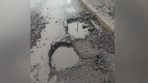 Matt Hunton Two deep holes filled with murky brown rainwater sit close together near the kerb of a residential road. The surrounding tarmac is broken and crumbling, with loose chunks of asphalt scattered around the edges. A street lamp is faintly reflected in the standing water.
