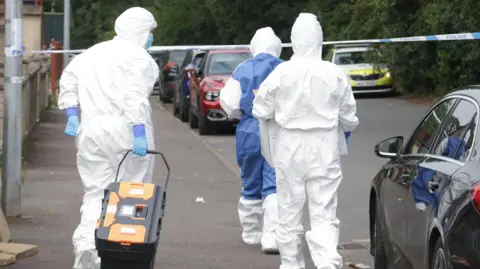 Pacemaker People in white hazmat suits walk down the street with their backs facing the camera. The street has cars parked up and a police car sits in the background.