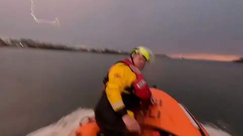 A man in an orange life boat is looking over his shoulder, towards the camera. He is wearing a yellow jacket and a red life jacket. The back ground is a mixture of water and cloudy skies with a faint tinge of orange as the sun sets.
