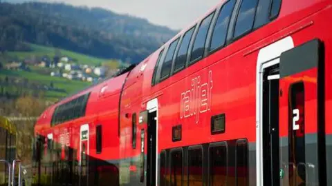 OBB An Austrian railjet train coloured in red at the top and grey at the bottom, seen with a background of a green grass-covered hilltop with a cluster of houses and trees further away