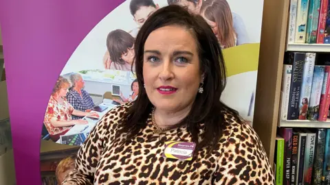 Patricia Curran has long brown hair and is wearing a leopard print top with a name tag spelling out 'Patricia'. She is standing in front of a Library poster and a bookcase full of books. 