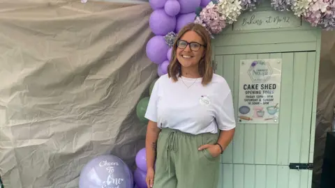 Sarah Day A women with short brunette hair. She is wearing a white top and green trousers whilst looking at the camera smiling. she is stood in front of a shed also green which is used to store her baked goods.