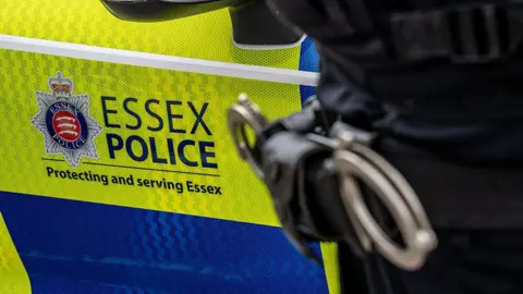 A close-up shot showing the side of an Essex Police car. The Essex Police logo can be seen above the words 'Protecting and serving Essex'. Handcuffs, which are blurred/out of focus, are also in the image.