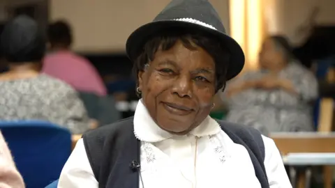 A black woman smiles into the camera, she is wearing a white blouse, black waistcoat and matching trilby hat