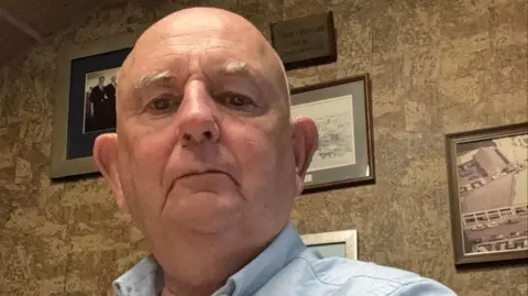 Nick Jenkins A bald man wearing a blue shirt is in room with several framed photos on the wall behind him.