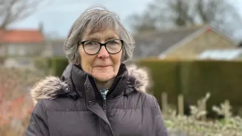 Helen Chamberlain is a woman in her 60s. She is wearing a brown coat with a fur lined hood and has grey hair and black-rimmed glasses.