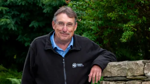 Cumbria Wildlife Trust Steve Trotter, chief executive of Cumbria Wildlife Trust. He has short salt and pepper hair and wears glasses. He is wearing a dark fleece over a blue shirt. His arm is leaning on a drystone wall and there are green trees behind him.