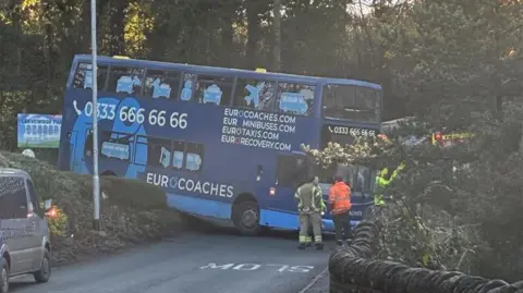 A blue double decker bus with a number along the top and the words "Eurocoaches, Eurominibuses, Eurotaxis" on the side. Its front end is blocking a narrow road, which bends to the right behind it. The back of the bus appears to have reversed over a wall/hedge off to the side of the road and is tilted upwards. There are firefighters standing near the front of the vehicle.