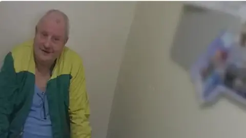 Steve Wright, wearing a yellow green jacket over a light blue top, sits down, looking in the general direction of the camera. He is in a plain room with pictures pinned to the wall.
