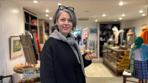 Bella Farrell stands inside a charity shop, which is warmly lit, with shelves and racks displaying a mix of clothing, accessories, and decorative items.
She is wearing a dark coat with a light grey scarf tucked inside it. She has shorter grey hair and a pair of glasses resting on top of their head.