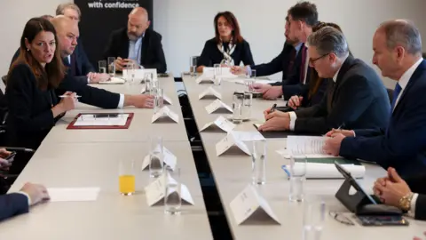 Reuters Ministers sit round a white table. They wear suits. There are name cards, paper, clipboards and glasses of water on the table.