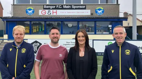BBC (LR) Gary Shufflebottom, Jordan Connolly, Melissa Gibbons y Stephen McCullough. Gary y Stephen usan sobre palabras de Navy Bangor FC, Jordan está en una camisa Adidas Rose y White, y Melissa está vestida con un traje negro. Los cuatro están parados uno al lado del otro en un campo de fútbol, fuera de un gran edificio de ladrillos que exhibe la cresta y patrocinadores de Bangor FC.