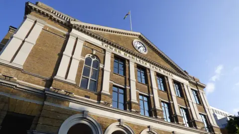LDRS An exterior view of the Tower Hamlets Town Hall, looking up at the front facade, with a clock and flagpole at the top.