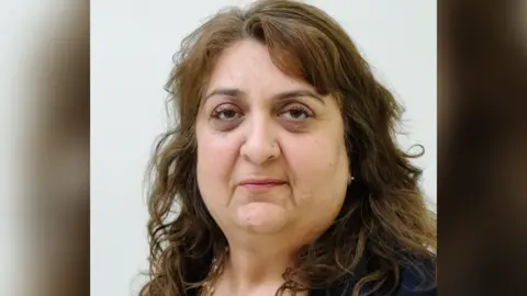 Puja Bedi is photographed in front a cream screen. She is expressionless in her councillor profile picture. She has shoulder length brown hair, studded earrings and a stud in her left nostril.
