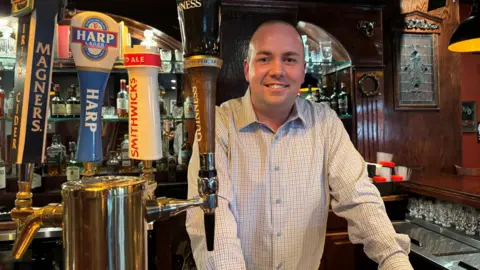 BBC Chris Devenny stands behind the beer taps of a pub, he is bald and is smiling and wearing a light-coloured small check shirt.