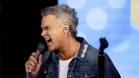 Robbie Williams photographed in the BBC Radio 1 Live Lounge. He is singing into a microphone and wearig a denim jacket with pearl flower design on collar