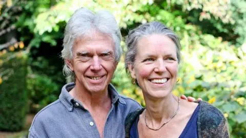 Picture of lady and her husband smiling. The husband has his hand on his wife's shoulder.They are both wearing blue. Both have grey hair. Behind them are trees and leaves. 