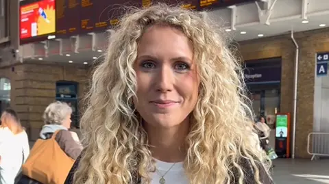 Georgia Pasco A woman in the middle of a train station. She's standing close to the camera and staring straight into the lens
