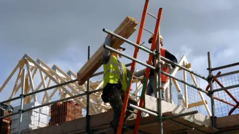 A pair of roofers work on a housing construction site. The timber structure of a roof can be seen and one man is holding planks on his shoulder and talking to the other man. Scaffolding, ladders and railings are being used.