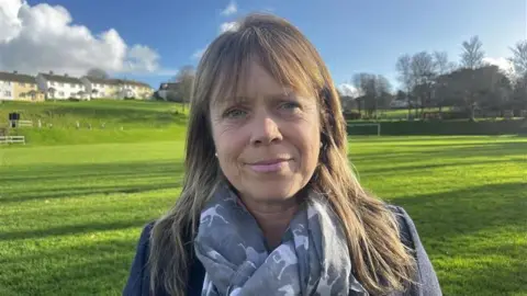 Dr Blunden is standing in a field smiling at the camera on a sunny day. She has long dark blonde hair and is wearing a coat and scarf.