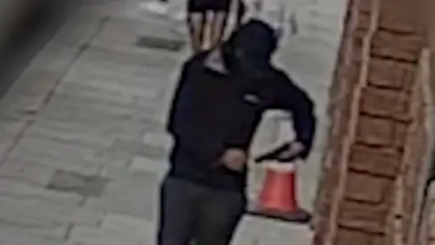 A hooded man dressed in black carries a gun in the street