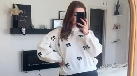 A woman in a white jumper with bows on it looks in the mirror holding her phone
