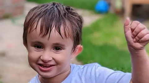Supplied A headshot of a young boy wearing a T-shirt while smiling and holding his hand up. He is looking directly into the camera.