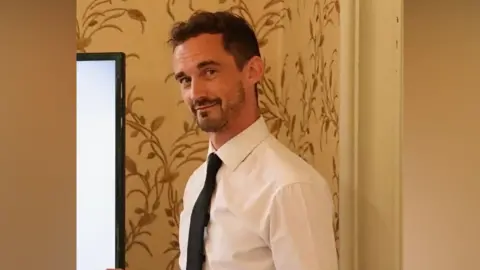 A photograph of a man looking at the camera wearing a white shirt and black tie 