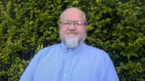 An image of Liberal Democrat councillor David Whipp. He's wearing a blue shirt and standing in front of a green leafy bush. He is wearing his hair in a ponytail, and has black rimmed glasses on. He is smiling.