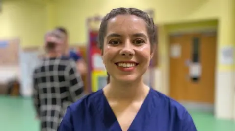 Isabella Sime, a woman with brown hair and a bright smile. She wears a blue dentist's uniform as she stands in a school corridor.