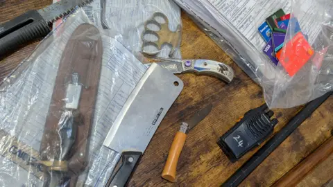 A selection of weapons which have been removed from loan sharks when the Illegal Lending Money Team have carried out raids on suspects' homes. The weapons on the table include a bronze knuckle duster, a meat cleaver, two flick knives, a hunting knife, a garden saw, a crow bar, a samurai sword and multiple bank cards which belong to victims.