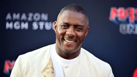 Getty Images Idris Elba wearing a cream jacket over a white t-shirt. He is at a red carpet event smiling directly at the camera
