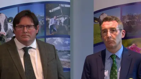 Two men. One is wearing glasses and a checked grey suit with a white shirt and tie. The other is wearing glasses and has a navy shirt on with a blue shirt and green tie.