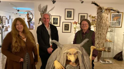 BBC Wendy Froud, Alan Lee, and Brain Froud stand amongst their art in the Green Hill Arts gallery. They are gathered around a large model of the Troll Witch, one of Wendy's creations. 