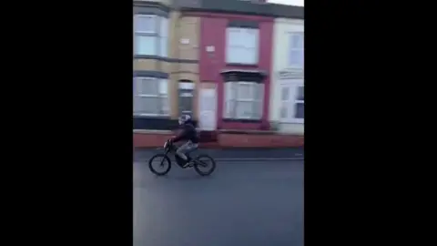 Boy on e-bike in police pursuit down street with terraced houses.