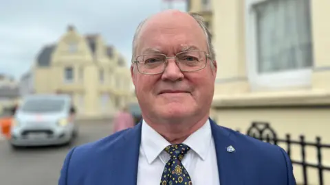 BBC A bald man with grey hair on the sides, wearing a blue suit, white shirt and patterned tie. He wears glasses and is standing in a residential street.