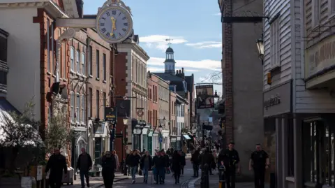 Rochester High Street on a sunny day, with many people walking along streets lined with historic brown and red brick buildings.