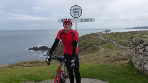 A man is wearing a red cycling top and black lyrca while holding a bike next to the sea and a sign for Lands End