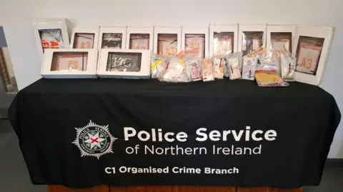 Image shows a black table, bearing the PSNI logo. A number of labelled white boxes are on top as well as clear bags, which show bank notes taken from the house search