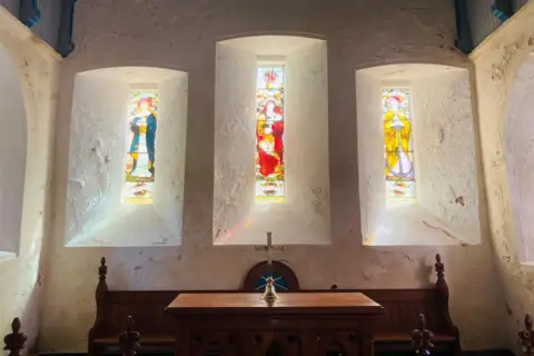 Three narrow stained glass windows in a church with light streaming through them.