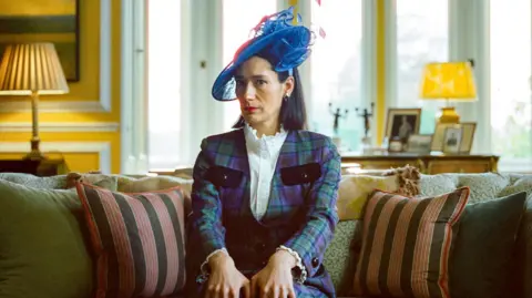 Metfilm Actor Sian Clifford wears a checked suit, a blue hat and sits on a sofa - adorned with cushions - in a drawing room. Lamps are behind her and on one table in the background are family photographs. The walls are painted yellow, with white panelling, and there is a large window.