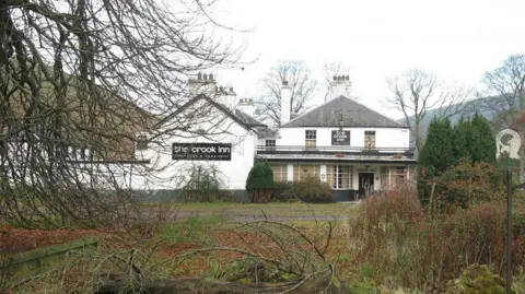 MJ Richardson The old Crook Inn site in Tweedsmuir with white walls and black signage viewed through trees