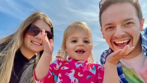 Family handout Rebecca Ableman (left), daughter Autumn (centre) and Chris Tuczemskyi (right) are all looking down at the camera on a sunny day with blue skies behind them. Rebecca has long blonde hair and sunglasses. Autumn is holding her hands up and is wearing a pink top with rainbows and unicorns on. She has blonde hair. Chris is also smiling at the camera and is wearing a blue checkered shirt with a white top. He has short brown hair and sunglasses on his head.
